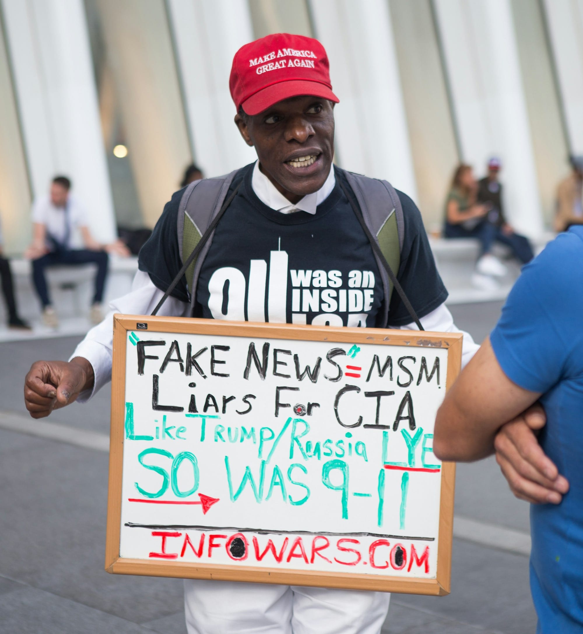 man in black and white shirt with red make America great again cap holding fake news=MSM sign near white building