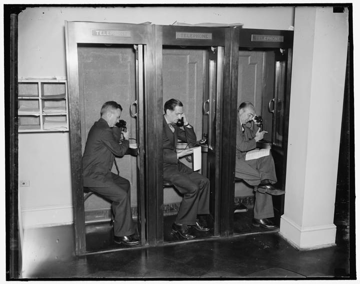 black and white photo of three male reporters seated and on the phone in three connected pay phone booths